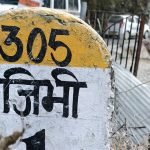 Jibhi travel guide: painted roadside marker reading Jibhi in Hindi, with village road and homes behind it.