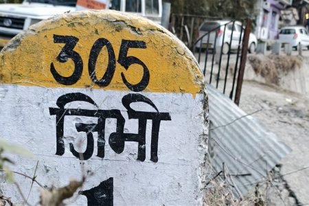 Jibhi travel guide: painted roadside marker reading Jibhi in Hindi, with village road and homes behind it.
