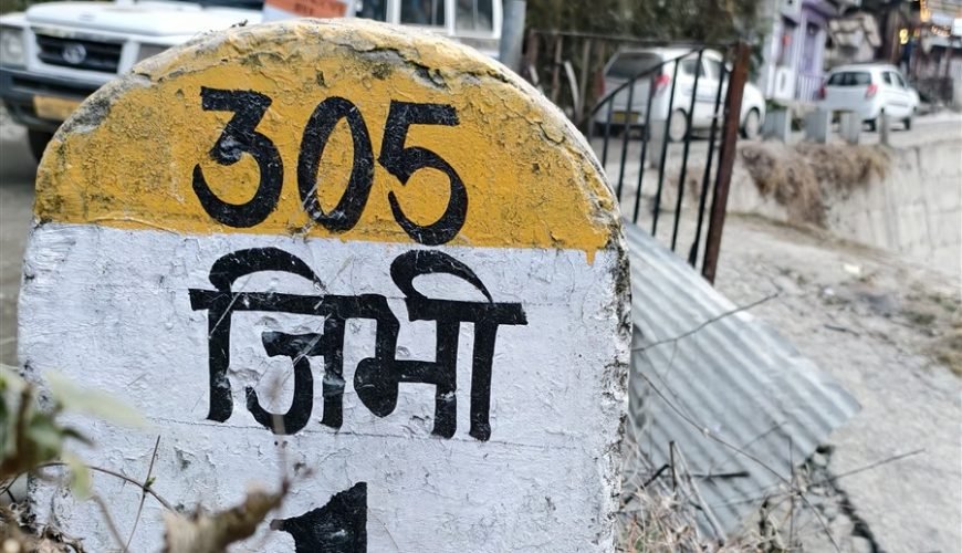 Jibhi travel guide: painted roadside marker reading Jibhi in Hindi, with village road and homes behind it.