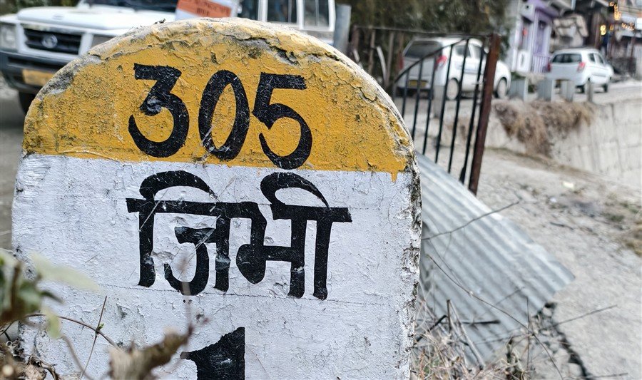 Jibhi travel guide: painted roadside marker reading Jibhi in Hindi, with village road and homes behind it.